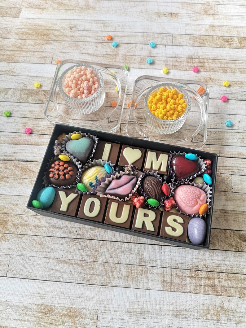 May include: A box of assorted chocolates arranged to spell "I love you". The chocolates are in a variety of shapes and colors, including hearts, lips, and letters. The box is surrounded by small, colorful sprinkles.