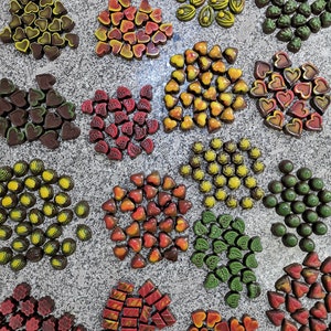 May include: Assortment of gourmet chocolates arranged in various shapes and colors, including hearts, leaves, stars, and swirls. The chocolates are displayed on a light gray surface.