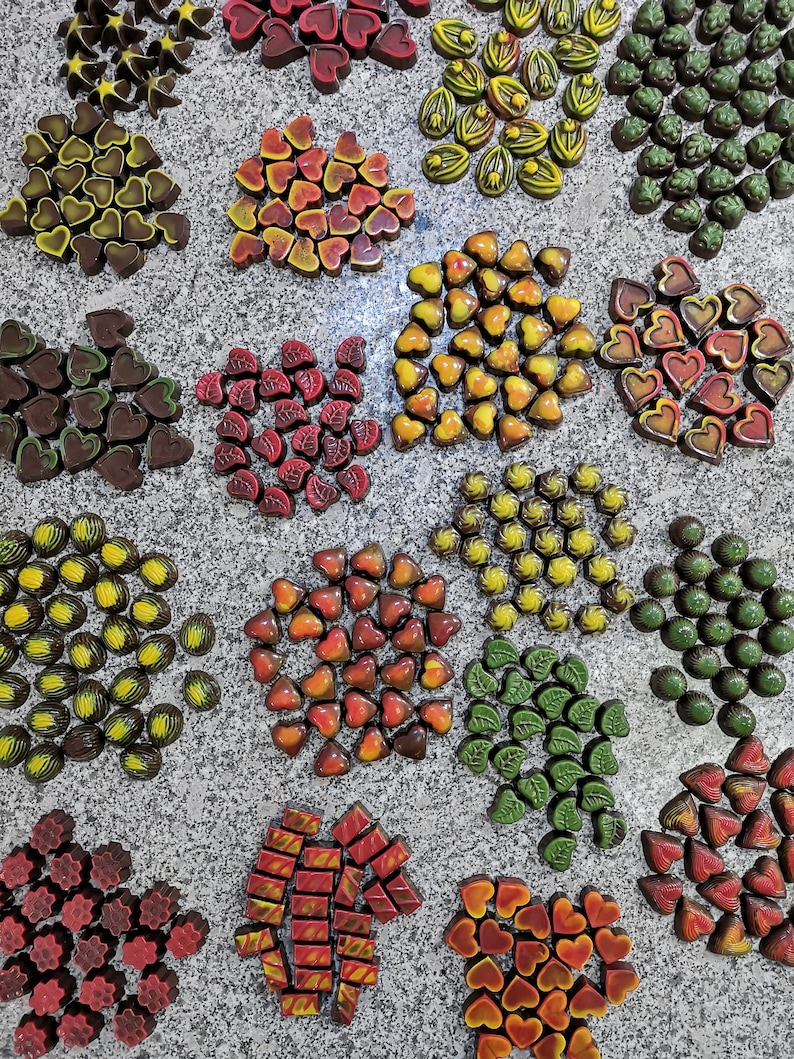 May include: Assortment of gourmet chocolates arranged in various shapes and colors, including hearts, leaves, stars, and swirls. The chocolates are displayed on a light gray surface.