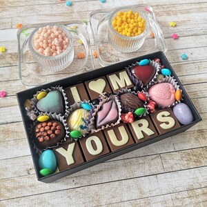 May include: A box of assorted chocolates arranged to spell "I LOVE YOU" with a heart-shaped chocolate in the center. The chocolates are in a black box with a white lid.