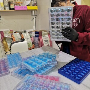 May include: A person wearing black gloves holds a clear plastic mold filled with small, dark gray chocolate pieces. The mold has 24 individual compartments.  Several other clear plastic molds are on a white marble surface. The molds are for making chocolate candies in various shapes, including hearts and capsules.