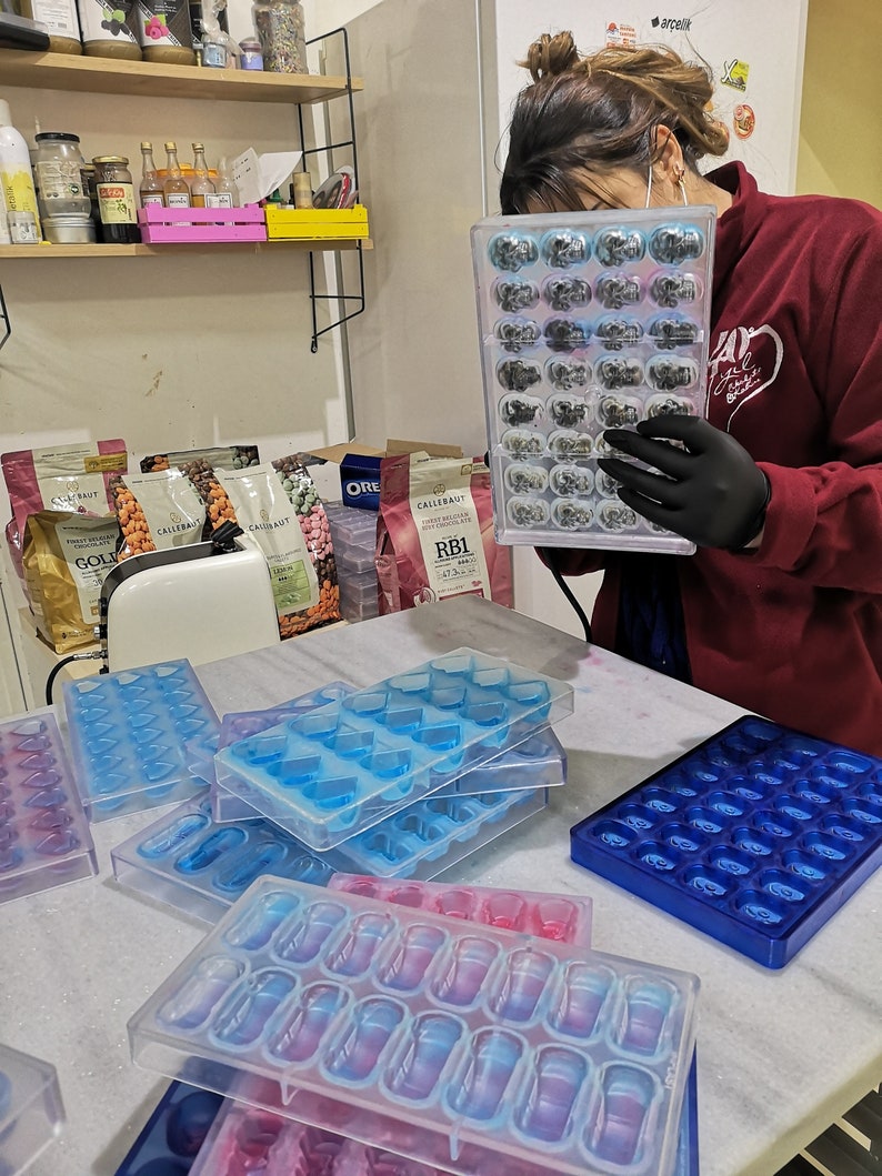 May include: A person wearing black gloves holds a clear plastic mold filled with small, dark gray chocolate pieces. The mold has 24 individual compartments.  Several other clear plastic molds are on a white marble surface. The molds are for making chocolate candies in various shapes, including hearts and capsules.