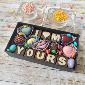 May include: A black box filled with chocolate candies arranged to spell "I love you". The box is on a wooden surface with colorful sprinkles scattered around it.