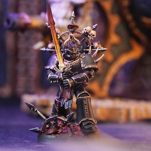 Puede incluir: Una figura en miniatura pintada de negro y dorado de un marine espacial sosteniendo una espada naranja brillante. La figura lleva una armadura negra con detalles dorados y está de pie sobre una base con una calavera y una rueda.