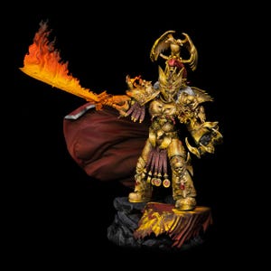 May include: A detailed gold-colored warrior figurine in ornate armor with a large eagle crest on the helmet. The figure holds a flaming orange sword and wears a flowing dark red cape. It stands on a rocky base featuring a red and gold banner.