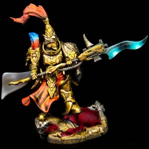 May include: A miniature warrior figure clad in ornate gold armour with a red plume and a blue-tipped feather. The figure holds a long polearm with a glowing blue blade and stands on a textured base featuring dark red fabric and metallic debris.