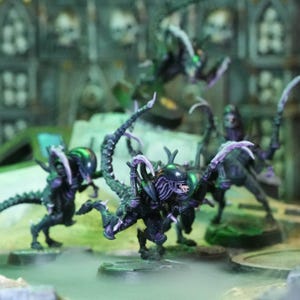 May include: A group of four black and purple alien figures with long, spindly legs and arms. The figures are posed in a dynamic, action-packed stance, suggesting a scene of combat or pursuit. The figures are painted with a detailed, realistic style, highlighting the intricate details of their anatomy.