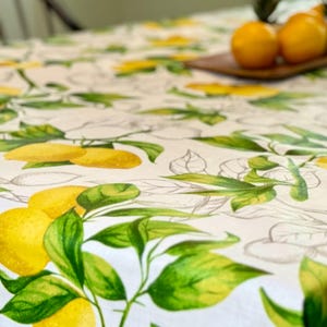 May include: A white tablecloth with a vibrant lemon and leaf pattern. The design features bright yellow lemons and green leaves, creating a fresh and summery aesthetic. The tablecloth is likely made of a durable fabric, perfect for kitchen or dining decor.