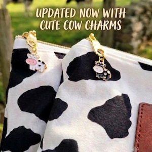 K&ouml;nnte beinhalten: Zwei wei&szlig;e Beutel mit schwarzem Kuhfleckenmuster, jeweils mit einem goldenen Rei&szlig;verschlussanh&auml;nger und einem kleinen Kuh-Charm aus Emaille. Der Text UPDATED NOW WITH CUTE COW CHARMS ist &uuml;ber den Beuteln zu sehen, die ein braunes Etikett haben.