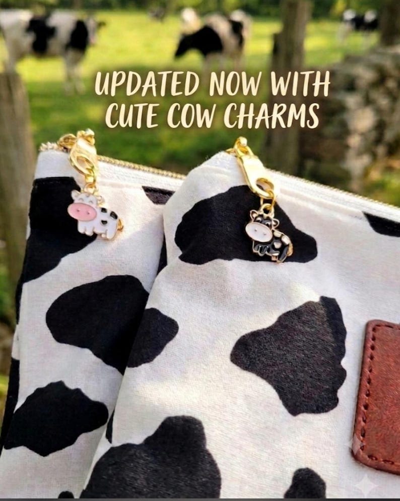 K&ouml;nnte beinhalten: Zwei wei&szlig;e Beutel mit schwarzem Kuhfleckenmuster, jeweils mit einem goldenen Rei&szlig;verschlussanh&auml;nger und einem kleinen Kuh-Charm aus Emaille. Der Text UPDATED NOW WITH CUTE COW CHARMS ist &uuml;ber den Beuteln zu sehen, die ein braunes Etikett haben.