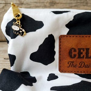 May include: A white fabric pouch with a black cow print pattern. The zipper features a gold-toned metal charm shaped like a cow. A rectangular brown leather-like patch is stitched to the front, displaying the text CEL and The Dai.