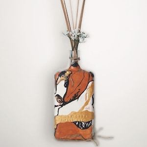 May include: A glass bottle vase with a painted orange, white and gold design. The vase is filled with dried reeds and a small bouquet of white flowers.