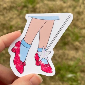 May include: A sticker featuring a pair of red glitter shoes with a blue sock and a star on the top of one shoe. The shoes are attached to a stick with a white star on the end.