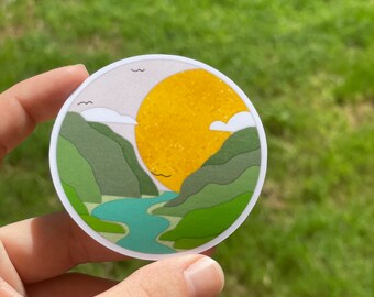 May include: A round sticker with a green mountain landscape and a yellow sun. The sun is shining brightly and there are two small white clouds in the sky. The sticker is perfect for adding a touch of nature to your belongings.