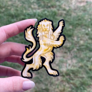 May include: A gold lion sticker with a black outline. The lion is standing on its hind legs with its front paws raised.