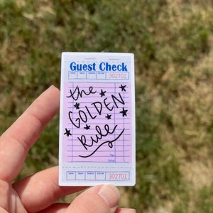 May include: A pink and white guest check sticker with the text "the GOLDEN rule" written in black cursive with stars around the words. The sticker is a replica of a restaurant guest check.