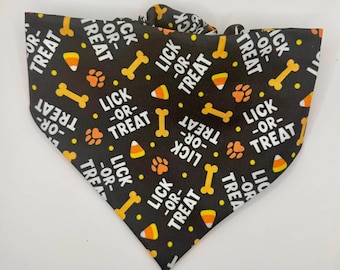 May include: A black triangular pet bandana featuring a repeating pattern of white text that reads LICK-OR-TREAT, interspersed with orange paw prints, yellow dog bones, small yellow dots, and orange, white, and yellow candy corn shapes.