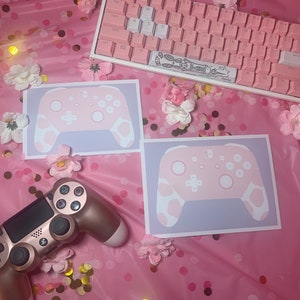 May include: A pink and white mechanical keyboard, two pink and white controller prints, and a gold controller are arranged on a pink surface with confetti and floral accents. The controller prints feature a cow-print design.