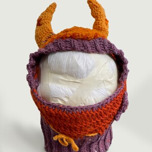 Könnte beinhalten: Eine lila und orangefarbene Häkelmütze mit Hörnern. Die Mütze hat eine spitze Kapuze und einen Bindfaden unten.