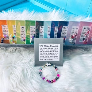 May include: A colorful beaded bracelet with a white cross charm, displayed with several packaged bracelets. The packages are arranged in a row, each with a different color scheme. A sign reads "The Messy Bracelet."