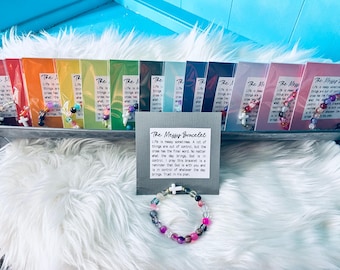 Op de afbeelding: Een kleurrijke armband met kralen en een witte kruisbedel, samen met verschillende verpakte armbanden. De pakketten zijn in een rij gerangschikt, elk met een ander kleurenschema. Een bord leest "The Messy Bracelet."