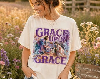 Op de afbeelding: Een crèmekleurig T-shirt met korte mouwen en een grafische print van een persoon in blauwe gewaden die bij een stenen waterput met een ander persoon praat. De tekst "GRACE UPON GRACE" staat in grote paarse letters boven en onder de afbeelding.