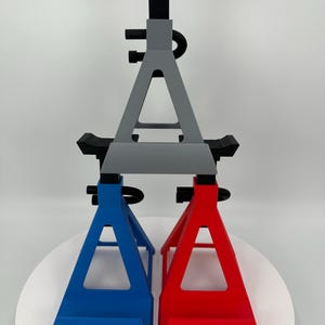 May include: A pair of automotive jack stands in blue and red, supporting a gray jack stand. The stands are made of a solid material and have a triangular base. The top of the stands have a black support.