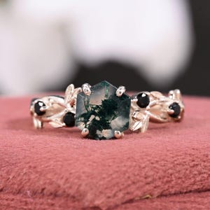 Pode incluir: Anel de prata com uma pedra de ágata musgosa hexagonal no centro. O anel apresenta detalhes de folhas e pedras pretas na banda. A ágata musgosa tem padrões verdes e brancos.