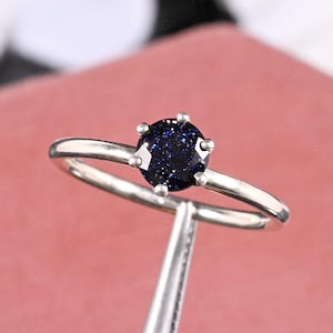 Pode incluir: Um anel de prata com uma pedra preciosa redonda azul escura com inclusões brilhantes. A pedra é engastada com seis garras. A banda é de prata lisa. O anel é mostrado sobre um fundo rosa pálido.