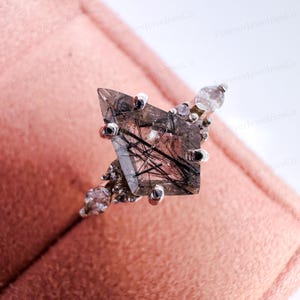 May include: A silver ring featuring a large, faceted, clear gemstone with black inclusions. The gemstone is set with small silver accents and smaller clear stones. The ring is displayed on a pink surface.