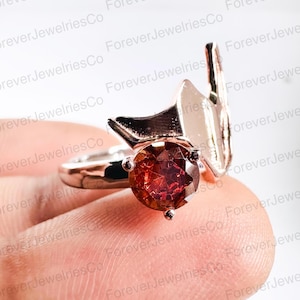 May include: A ring with a large, red gemstone set in a gold setting. The ring is designed with a unique, angular shape.