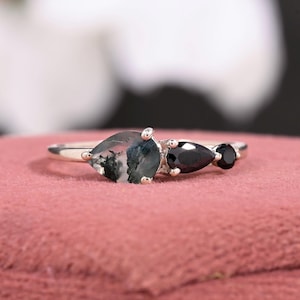 May include: A silver ring featuring a pear-shaped moss agate stone with green and white inclusions, flanked by two black onyx stones. The ring is set on a pink surface.