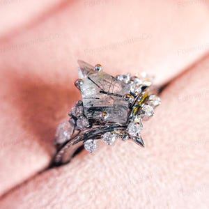 Könnte beinhalten: Ein silberner Ring mit einem großen, klaren, facettierten Edelstein mit schwarzen Einschlüssen. Der Edelstein ist in einer Krappenfassung gefasst und von kleineren, klaren Edelsteinen umgeben. Der Ring befindet sich auf einem rosafarbenen Hintergrund.