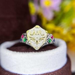 May include: A silver and gold ring with a shield-shaped design featuring a stylized bird with wings spread. The shield is encrusted with small clear stones and surrounded by four colored stones: red, green, blue, and pink.