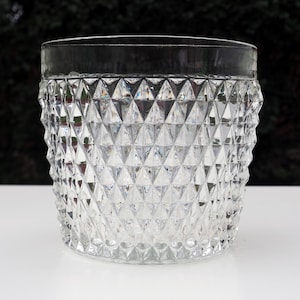 May include: A clear glass tumbler featuring a prominent diamond-cut geometric pattern across its exterior surface. The vessel has a smooth, unadorned rim and a cylindrical shape, showcasing a reflective, faceted texture throughout the body.
