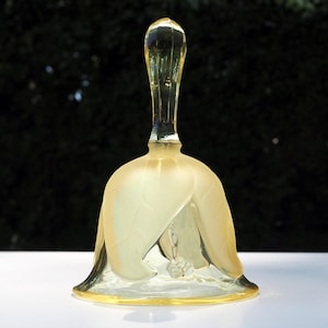 May include: A translucent yellow glass bell featuring a frosted finish on the exterior body, which is shaped like a flower with prominent leaf-like petals. The handle is smooth and clear, and a small clear glass clapper hangs inside the bell.