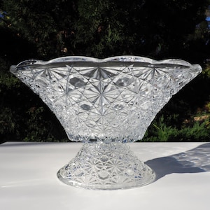 May include: A clear, cut-glass punch bowl with a pedestal base. The bowl has a scalloped rim and a diamond-cut pattern. The pedestal base also features a diamond-cut design. The bowl is sitting on a white surface.