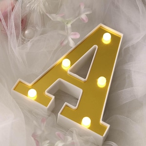 May include: A gold-colored, illuminated letter "A" with a white border. The letter is lit with small, round, warm-toned lights. The background is a soft, white, gauzy fabric with floral accents.