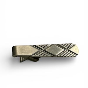 Puede incluir: Un pasador de corbata plateado con un patrón geométrico de diamantes y líneas. El pasador tiene forma rectangular con un extremo curvado y un cierre en la parte posterior. El diseño está grabado en el metal.