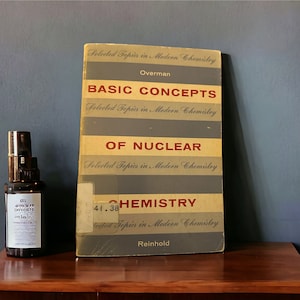 May include: A vintage book titled "Basic Concepts of Nuclear Chemistry" by Overman. The book cover is brown with gray stripes and the title is in red lettering. The book is sitting on a wooden table next to a clear glass bottle with a black cap.