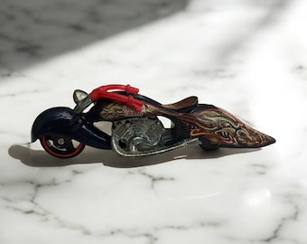 May include: A toy motorcycle with a dark blue body, red accents, and a brown seat. The motorcycle features intricate flame designs in shades of gold and red. The engine is silver, and the wheels have red rims.