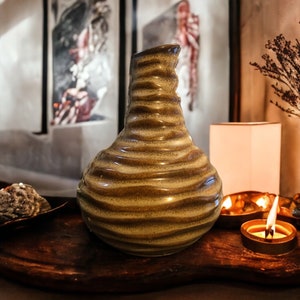 May include: A green and brown ceramic vase with a wavy design sits on a wooden tray with lit candles and decorative stones.