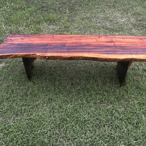 May include: A long, rustic wooden bench with a live edge top and dark brown stain. The bench has two simple black metal legs.