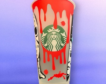 May include: A clear plastic reusable cup with a red, white, and black design. The design features a dripping blood effect, a Starbucks logo, and a horror movie character.