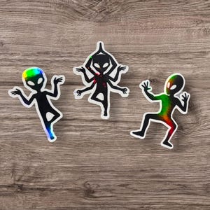 May include: Three holographic alien stickers in black with white outlines. One alien has rainbow coloring, another has a multi-armed design, and the third has rainbow coloring. The stickers are on a wooden surface.
