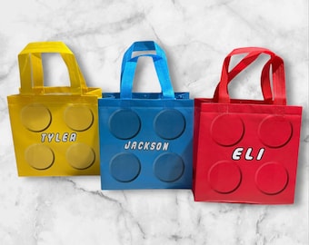 May include: Three reusable shopping bags with a Lego brick design. The bags are yellow, blue, and red. Each bag has a name printed on it: Tyler, Jackson, and Eli.