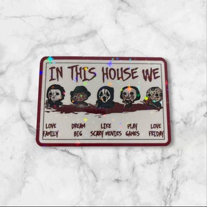 May include: A red and white sign with the text "In this house we" and a row of five horror movie characters. The text below the characters reads "Love family, Dream big, Like scary movies, Play games, Love Friday."
