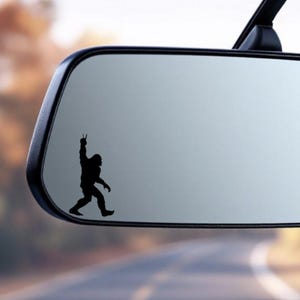 May include: A black silhouette of Bigfoot giving a peace sign, affixed to a car's rearview mirror. The image is a sticker, with the background showing a blurred outdoor scene. The Bigfoot figure is walking.