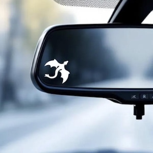 May include: A white dragon decal is affixed to a car's rearview mirror. The dragon is depicted in a stylized, minimalist design. The mirror is black and reflects a blurred outdoor scene.