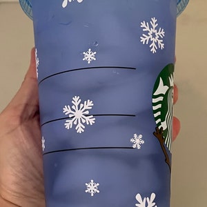 Puede incluir: Un vaso fr&iacute;o de pl&aacute;stico azul de Starbucks con tapa a juego y pajita verde. La taza est&aacute; decorada con copos de nieve blancos. El logotipo de Starbucks es visible en el lateral.